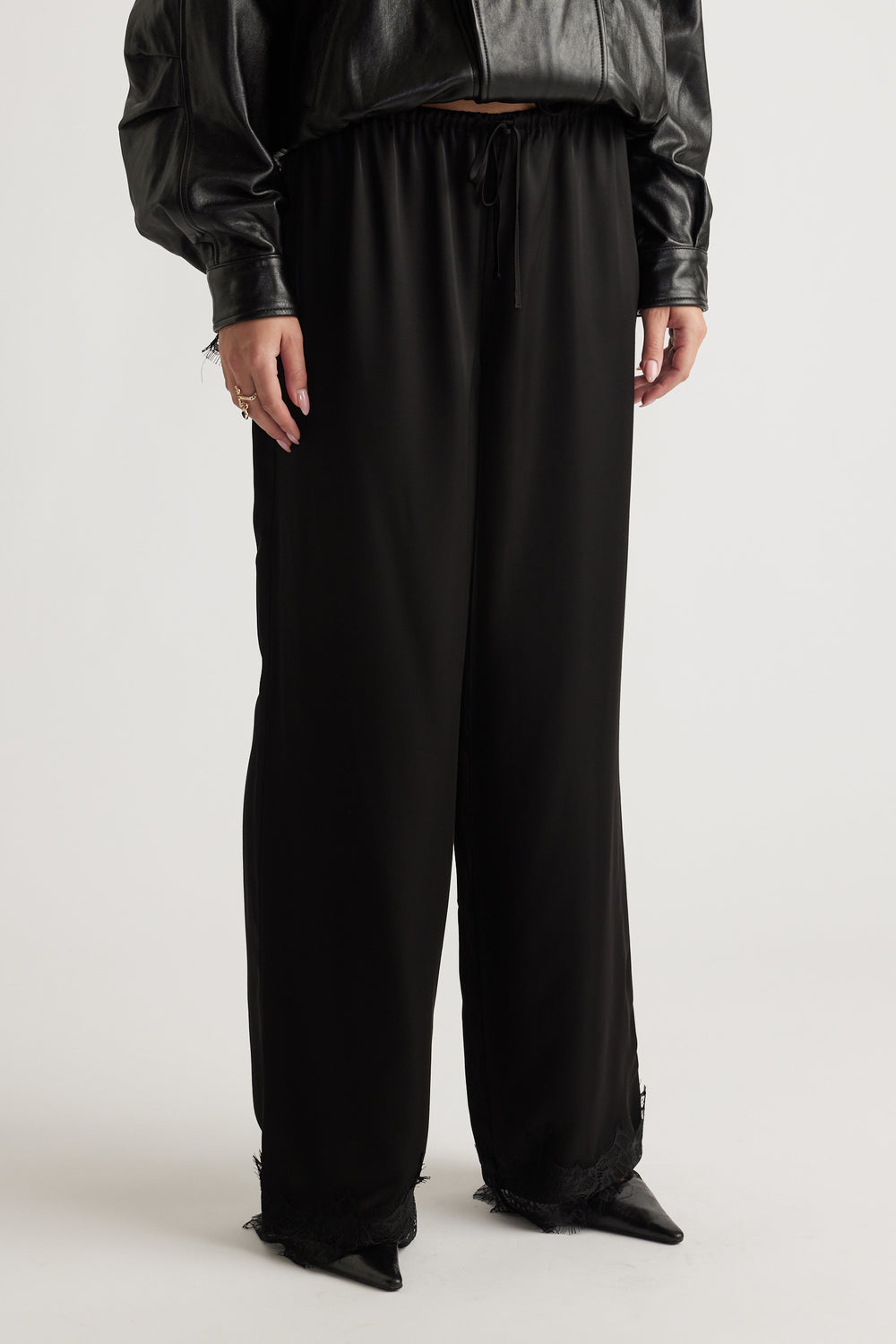 Black wide-leg satin pants with lace trim hem, elastic waistband, and drawstring, styled with a cropped leather jacket.