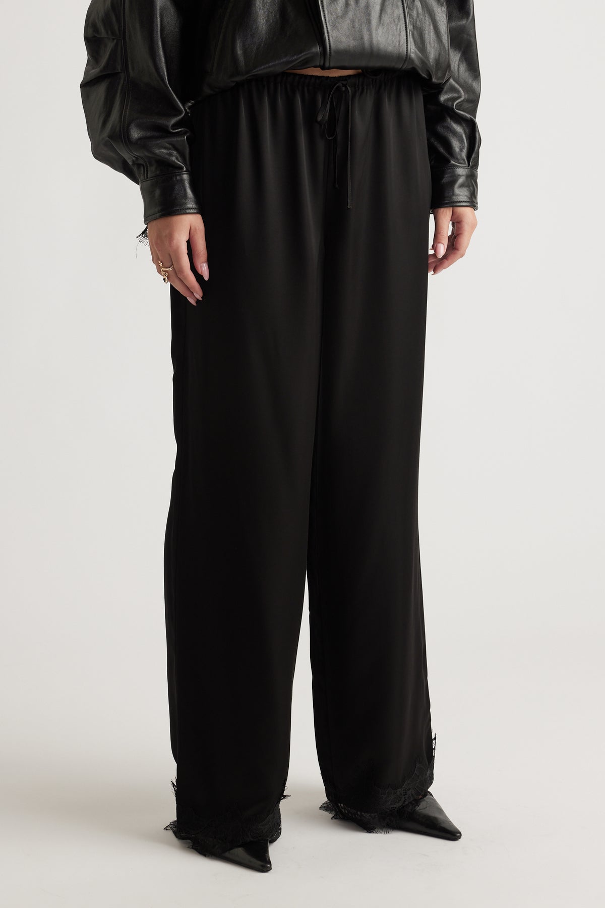 Black wide-leg satin pants with lace trim hem, elastic waistband, and drawstring, styled with a cropped leather jacket.