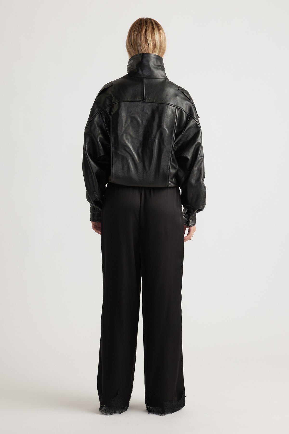 Woman seen from behind wearing loose black satin pants with lace trim at the hem and a cropped black leather jacket