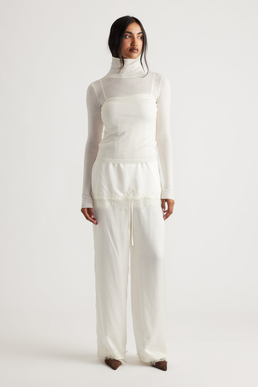 Woman wearing ivory satin pants with delicate lace trim paired with a sheer white turtleneck top and brown shoes