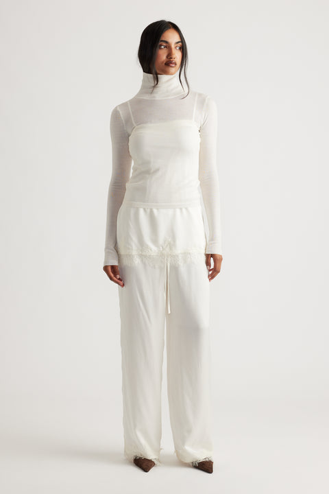 Woman wearing ivory satin pants with delicate lace trim paired with a sheer white turtleneck top and brown shoes