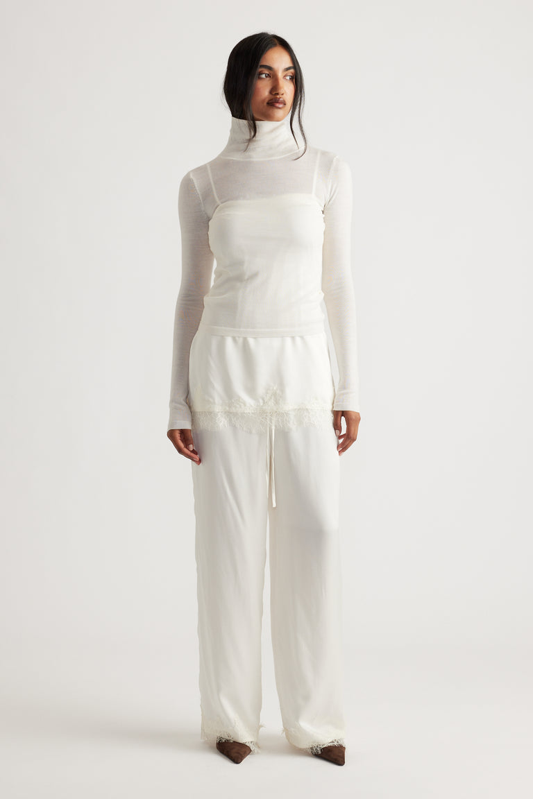 Woman wearing ivory satin pants with delicate lace trim paired with a sheer white turtleneck top and brown shoes