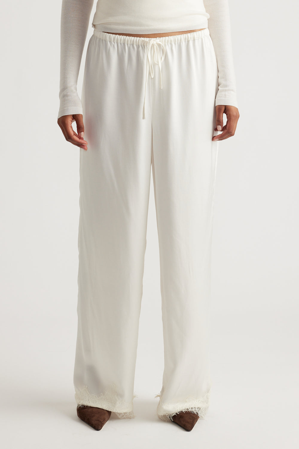 Front view of a woman wearing loose, ivory satin pants with delicate lace trim at the hem, styled with a fitted long-sleeve top