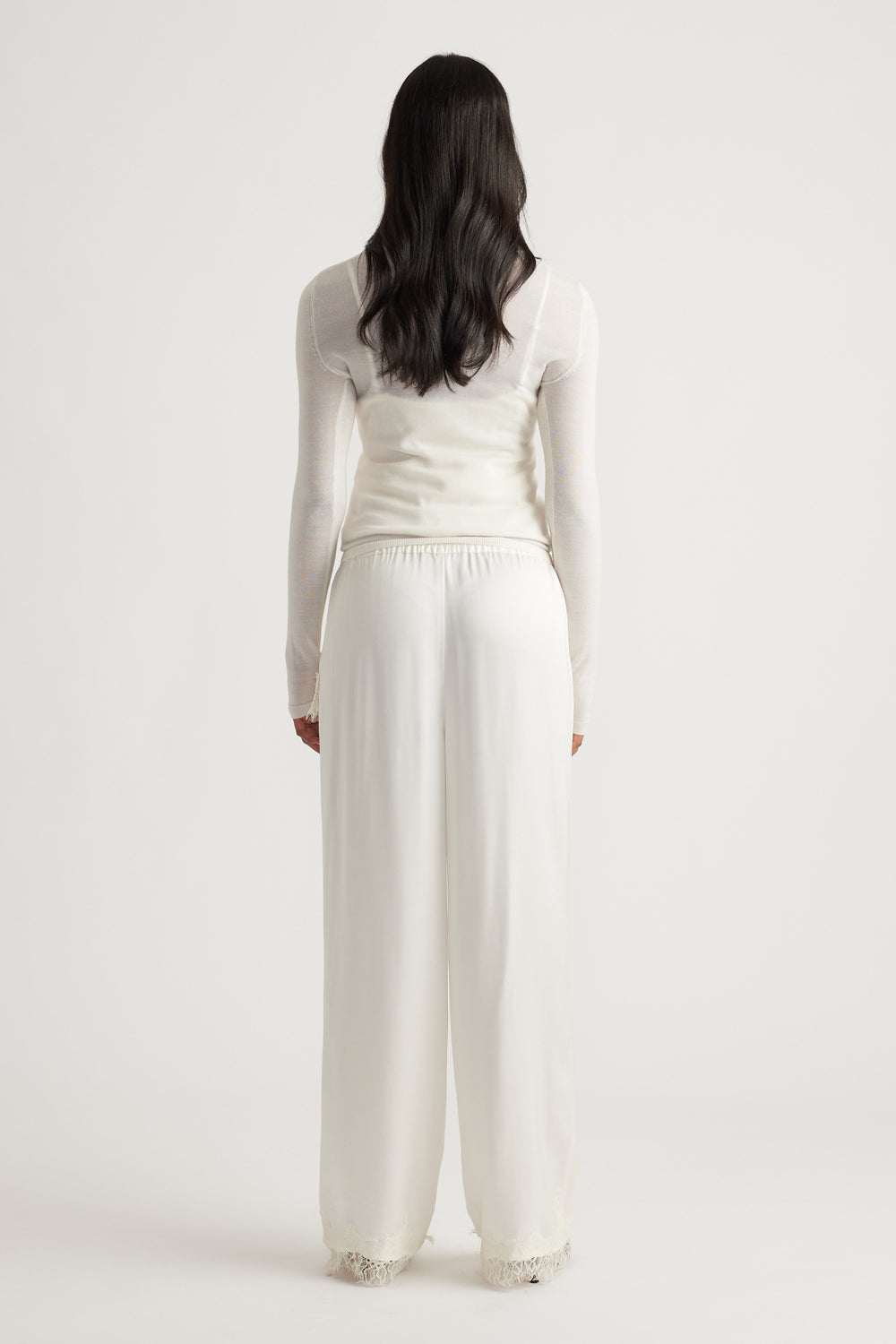 Woman wearing flowing ivory satin pants with delicate lace trim at the hem, paired with a fitted sheer long sleeve top.