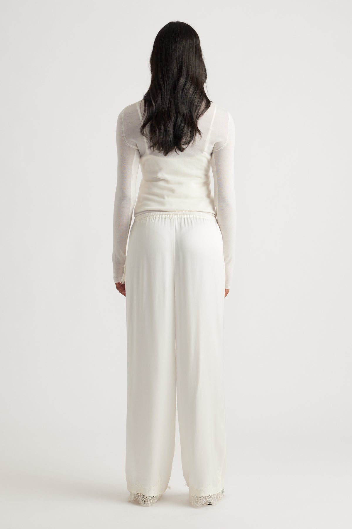 Woman wearing flowing ivory satin pants with delicate lace trim at the hem, paired with a fitted sheer long sleeve top.