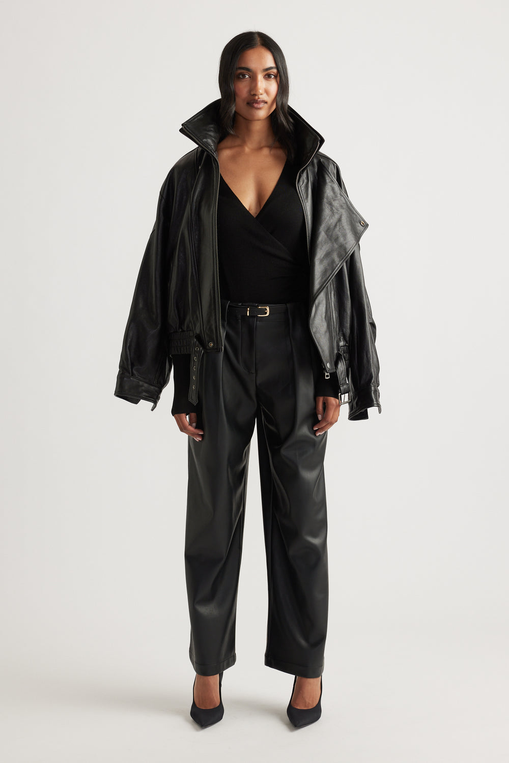 Woman wearing wide-leg black faux leather pants with pleats and a belted waist paired with a black wrap top and jacket