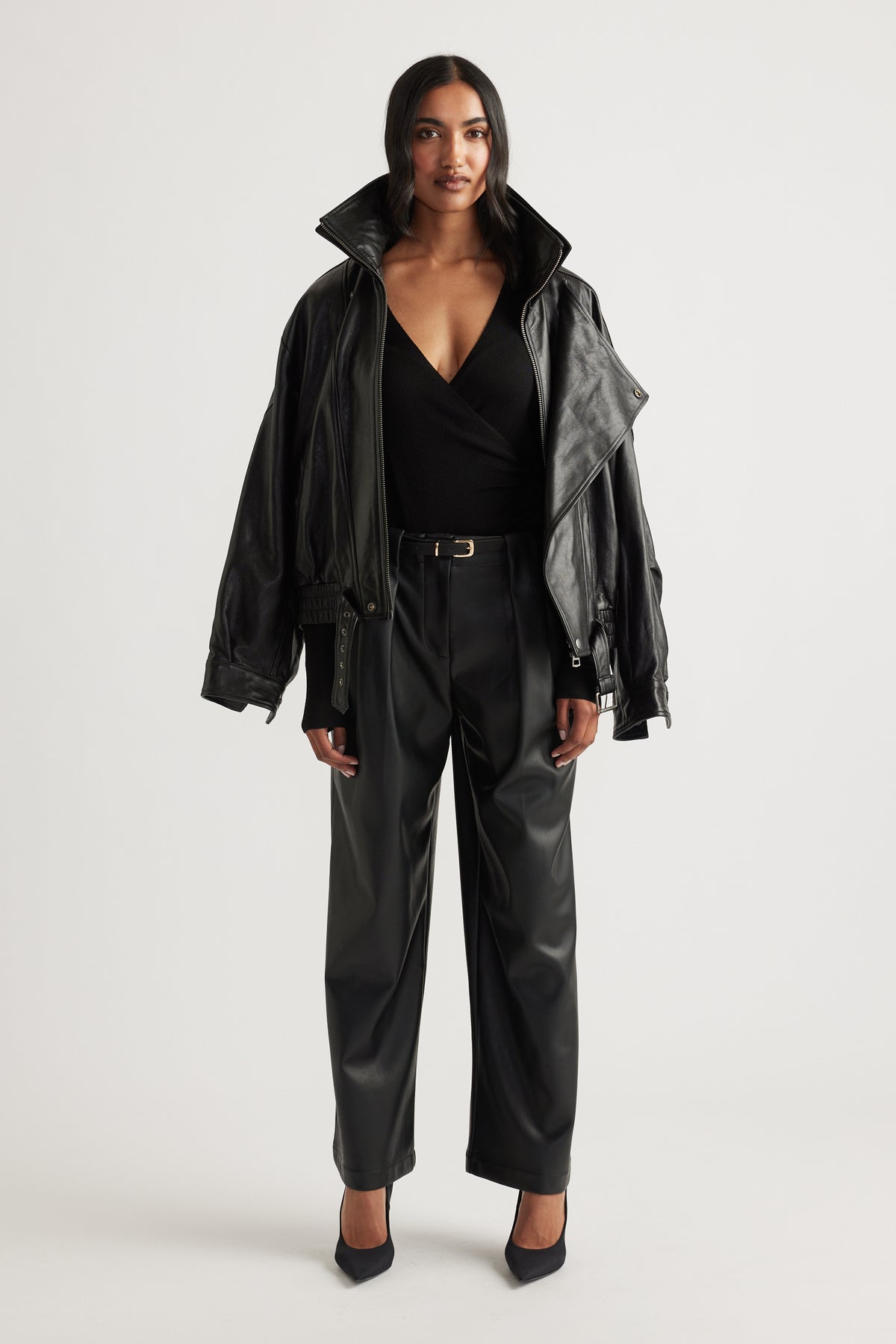 Woman wearing wide-leg black faux leather pants with pleats and a belted waist paired with a black wrap top and jacket