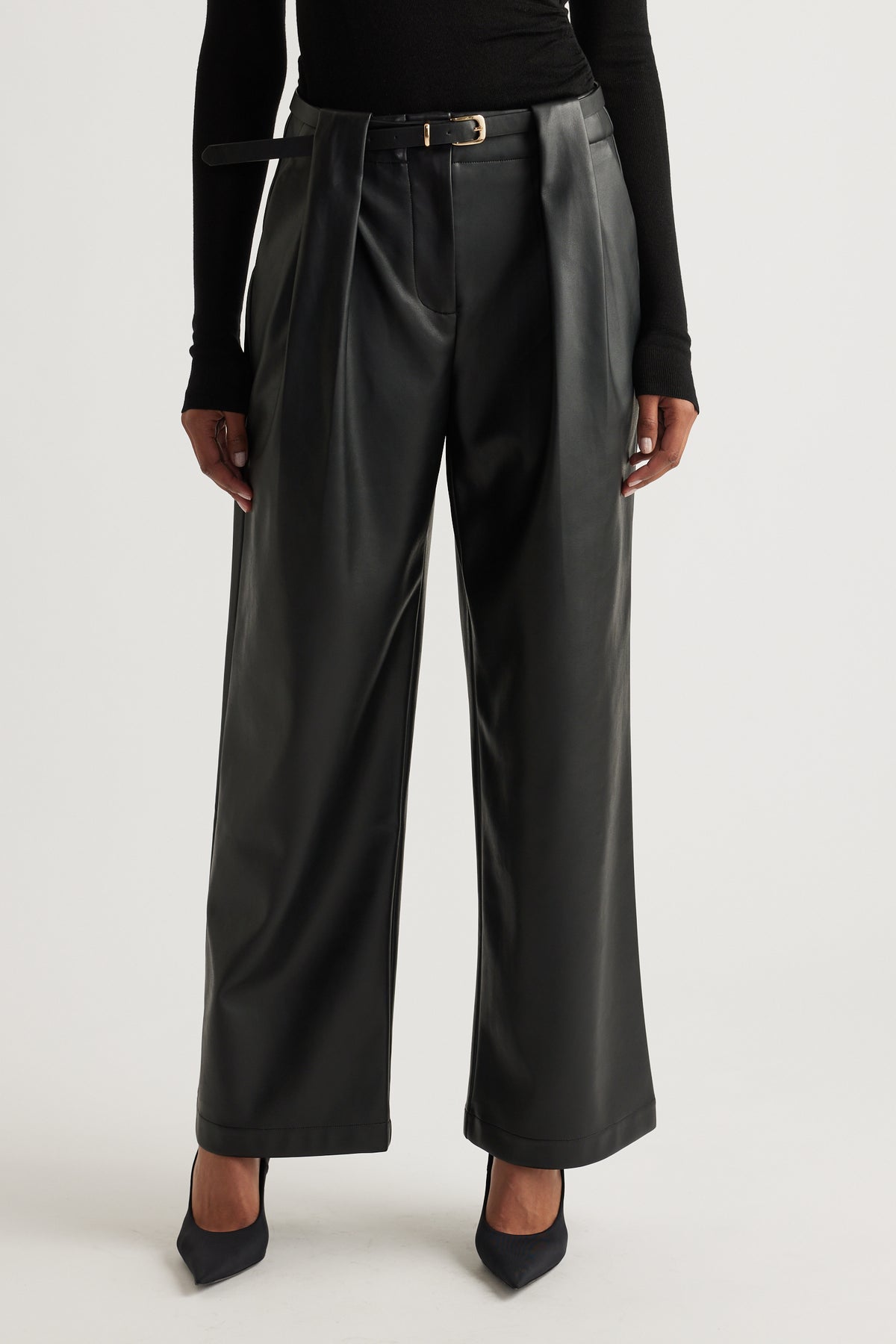 Front view of wide-leg black faux leather pants styled with a slim belt and paired with pointed black heels