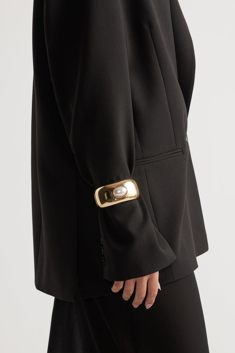 Monica Stone Cuff Bracelet in polished gold tone with central pearl accent, styled over black tailored blazer sleeve.