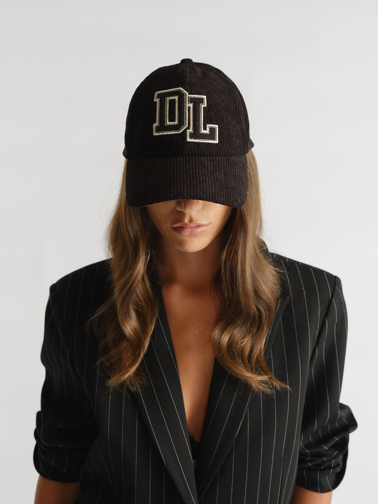 Woman wearing a black D-luxe Kim Cord Cap with "DL" embroidery, paired with a pinstripe blazer for stylish layering.