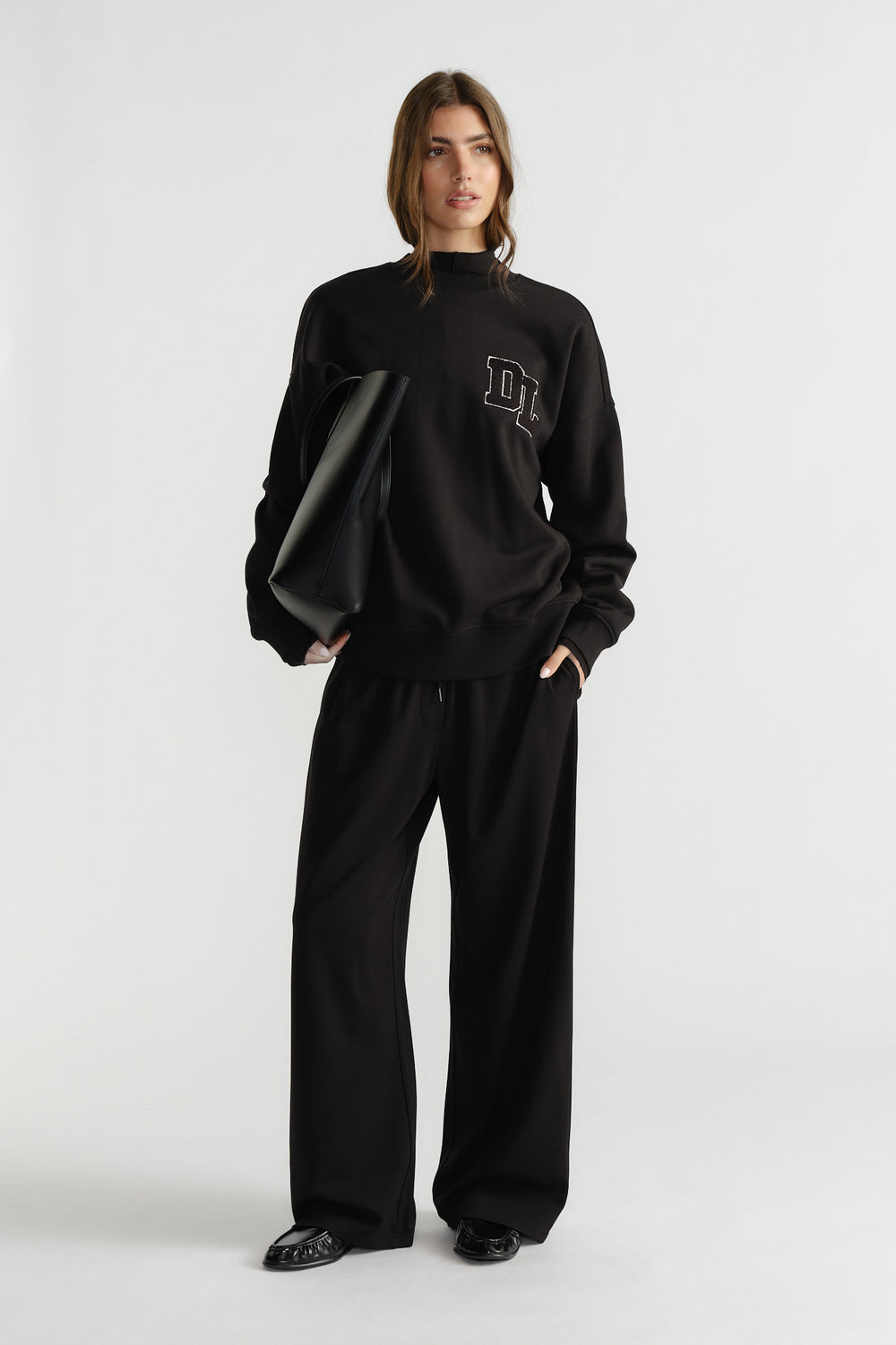 Woman wearing relaxed black crew sweat with logo detail, paired with loose black pants and black pointed shoes.