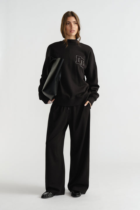 Woman wearing relaxed black crew sweat with logo detail, paired with loose black pants and black pointed shoes.