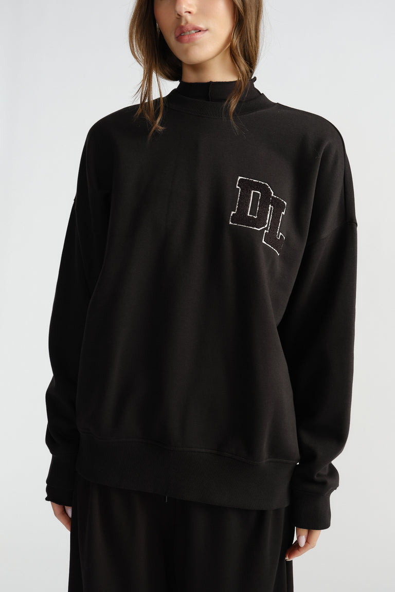 Woman wearing relaxed black crew neck sweatshirt with subtle embroidered logo on chest and long sleeves.