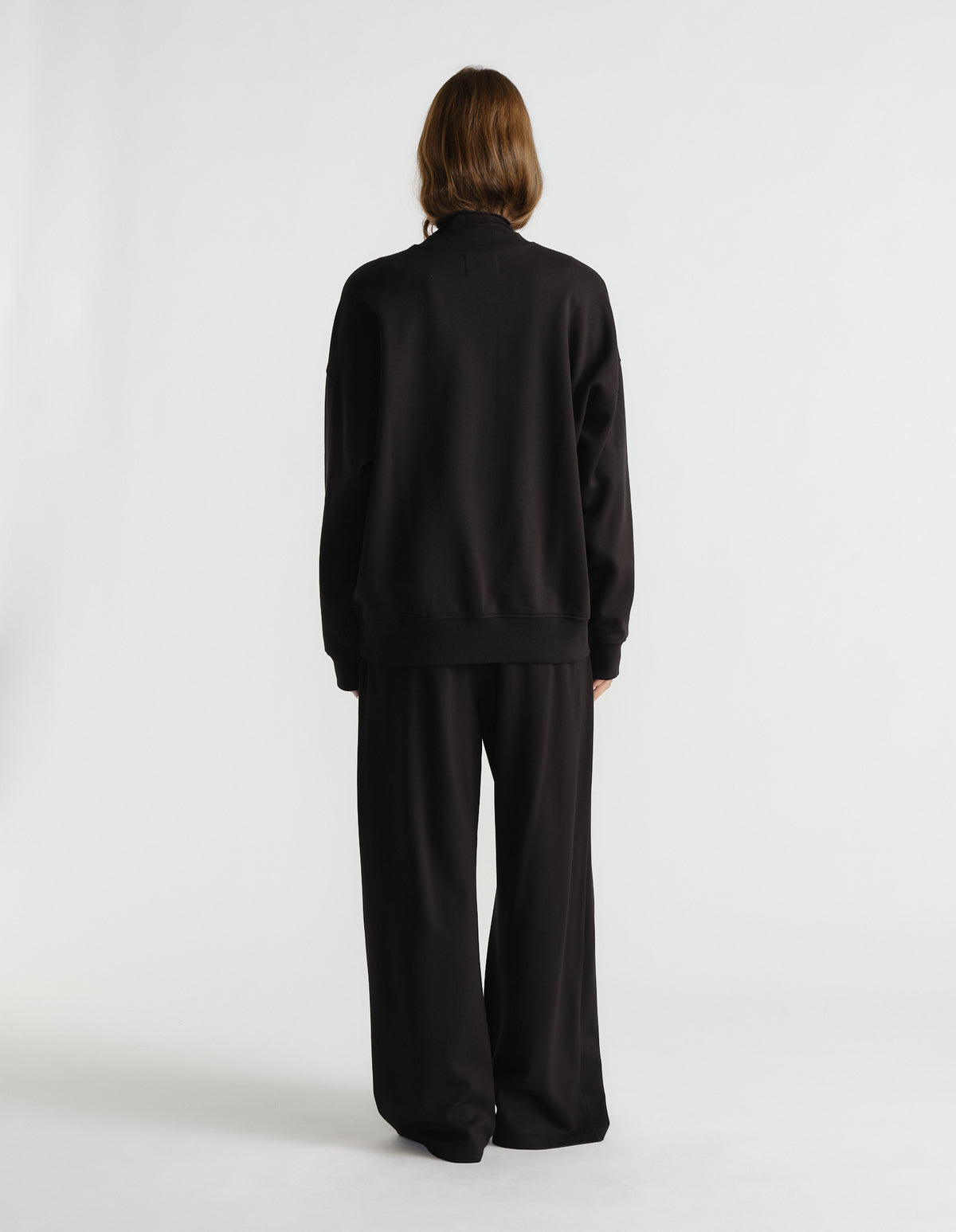 Back view of a woman wearing a relaxed black crew neck sweat paired with wide-leg black pants against a plain backdrop