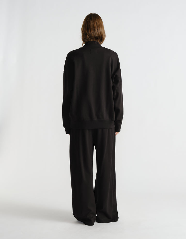 Back view of a woman wearing a relaxed black crew neck sweat paired with wide-leg black pants against a plain backdrop