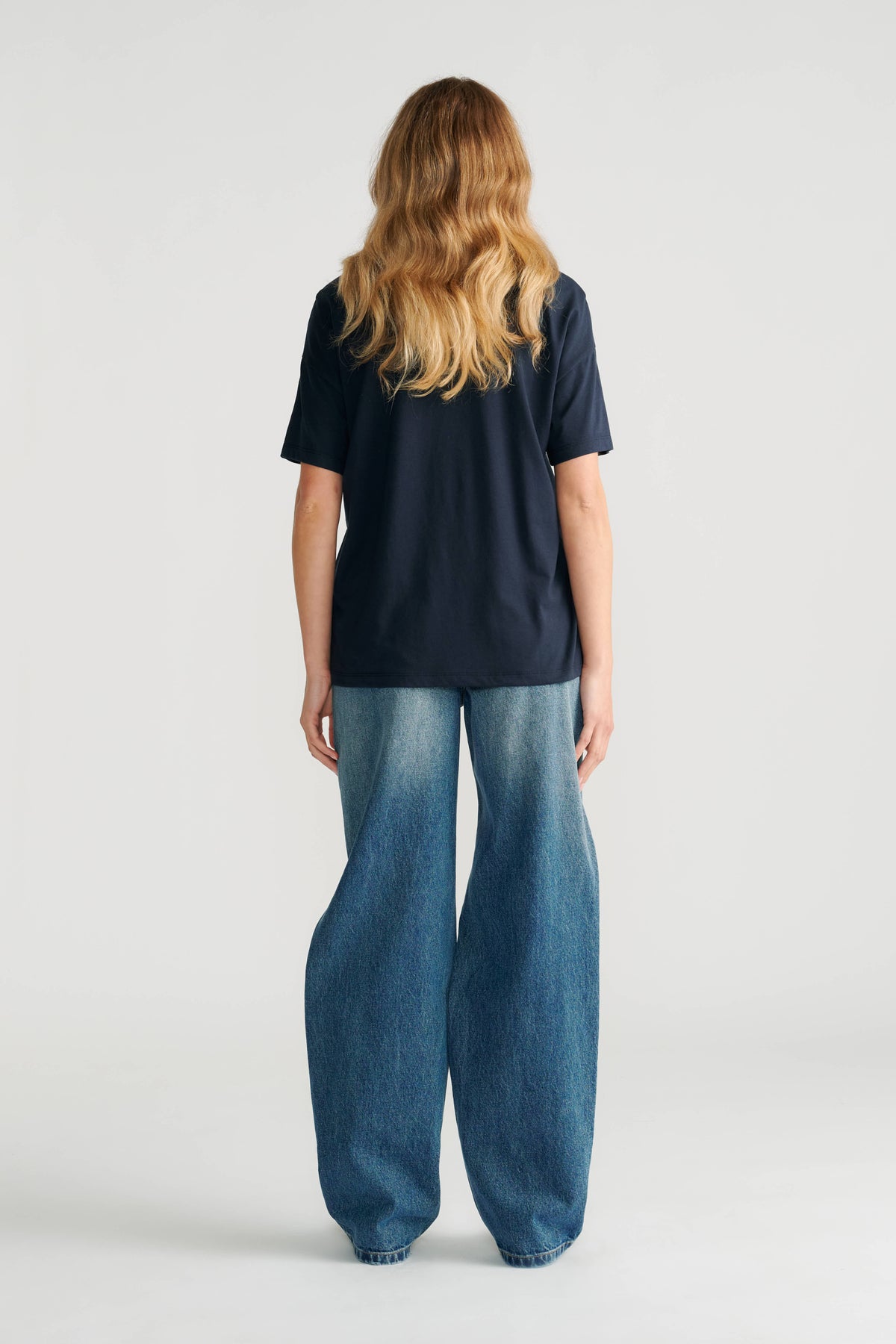 Woman wearing a classic navy short sleeve tee paired with relaxed wide-leg faded blue jeans shown from the back.