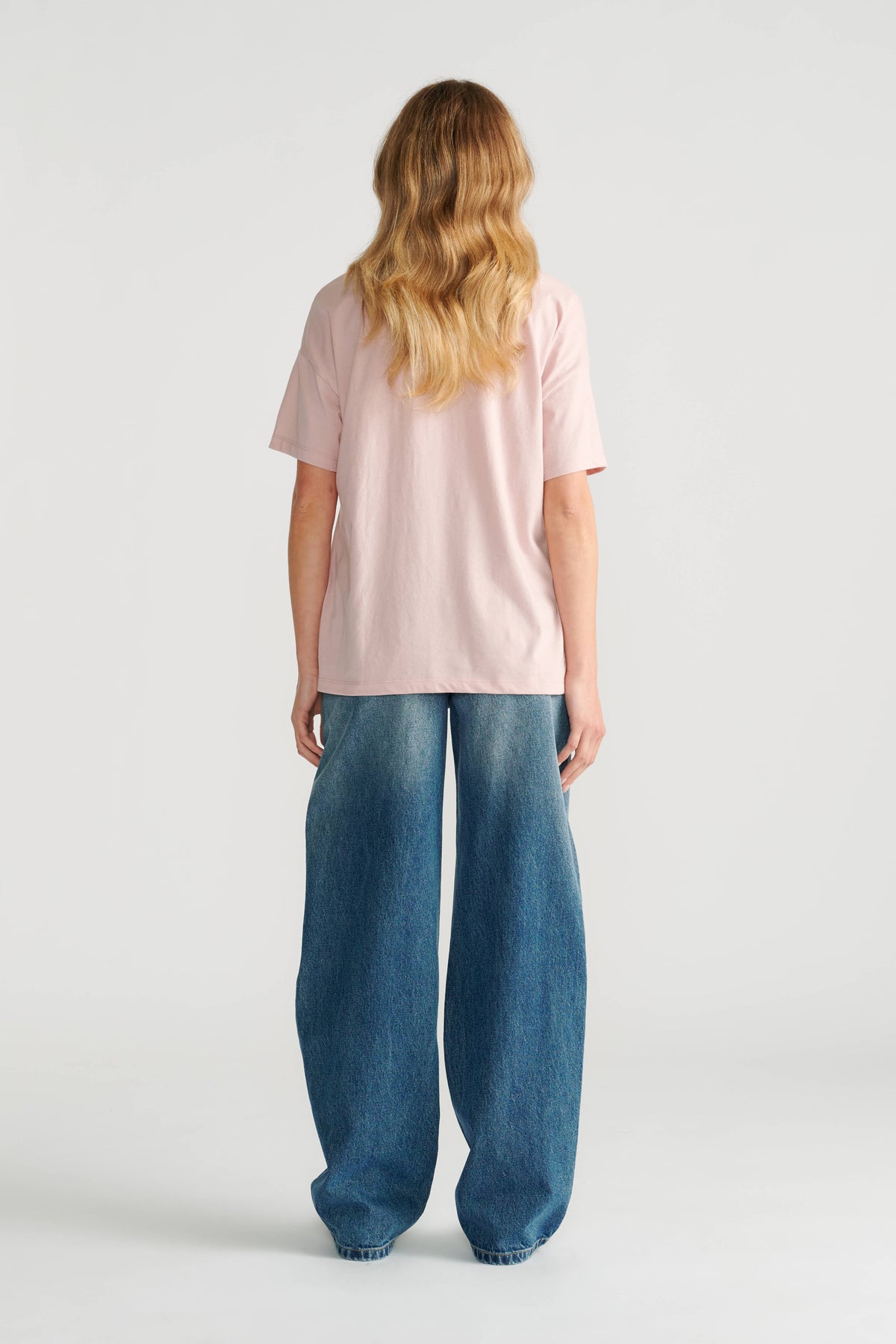 Woman wearing a soft pink short sleeve tee paired with loose, wide-leg blue denim jeans shown from behind