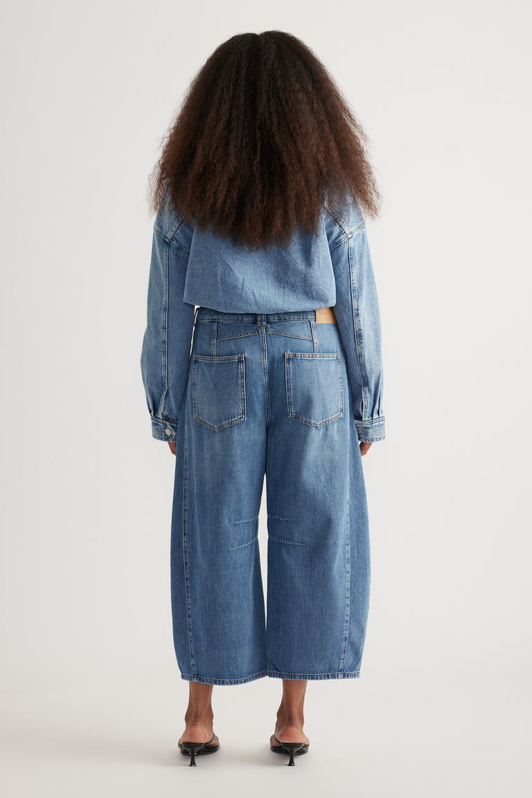 Back view of a woman wearing cropped blue barrel-leg jeans with a denim jacket and black heels
