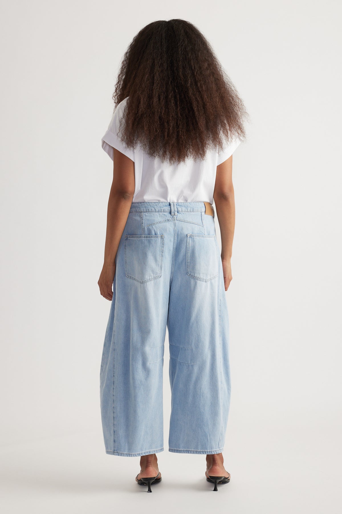Back view of woman wearing high rise cropped light blue barrel-leg jeans with a white tee and kitten heels