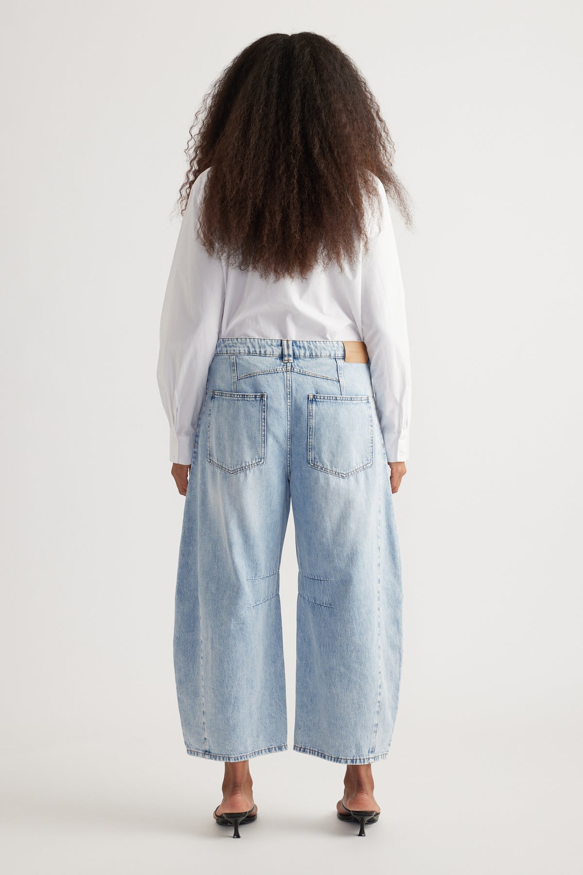 Back view of woman wearing light blue cropped barrel leg jeans with back pockets, a white blouse and heels