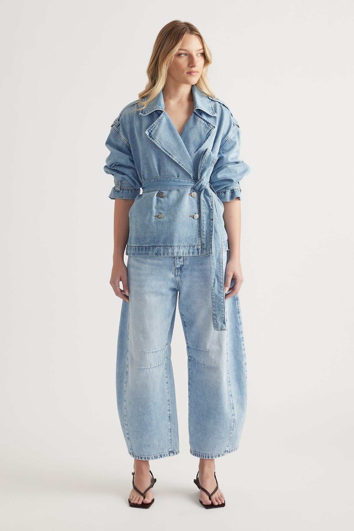 Woman wearing a light wash denim belted jacket paired with high-rise wide-leg denim jeans and black heeled sandals