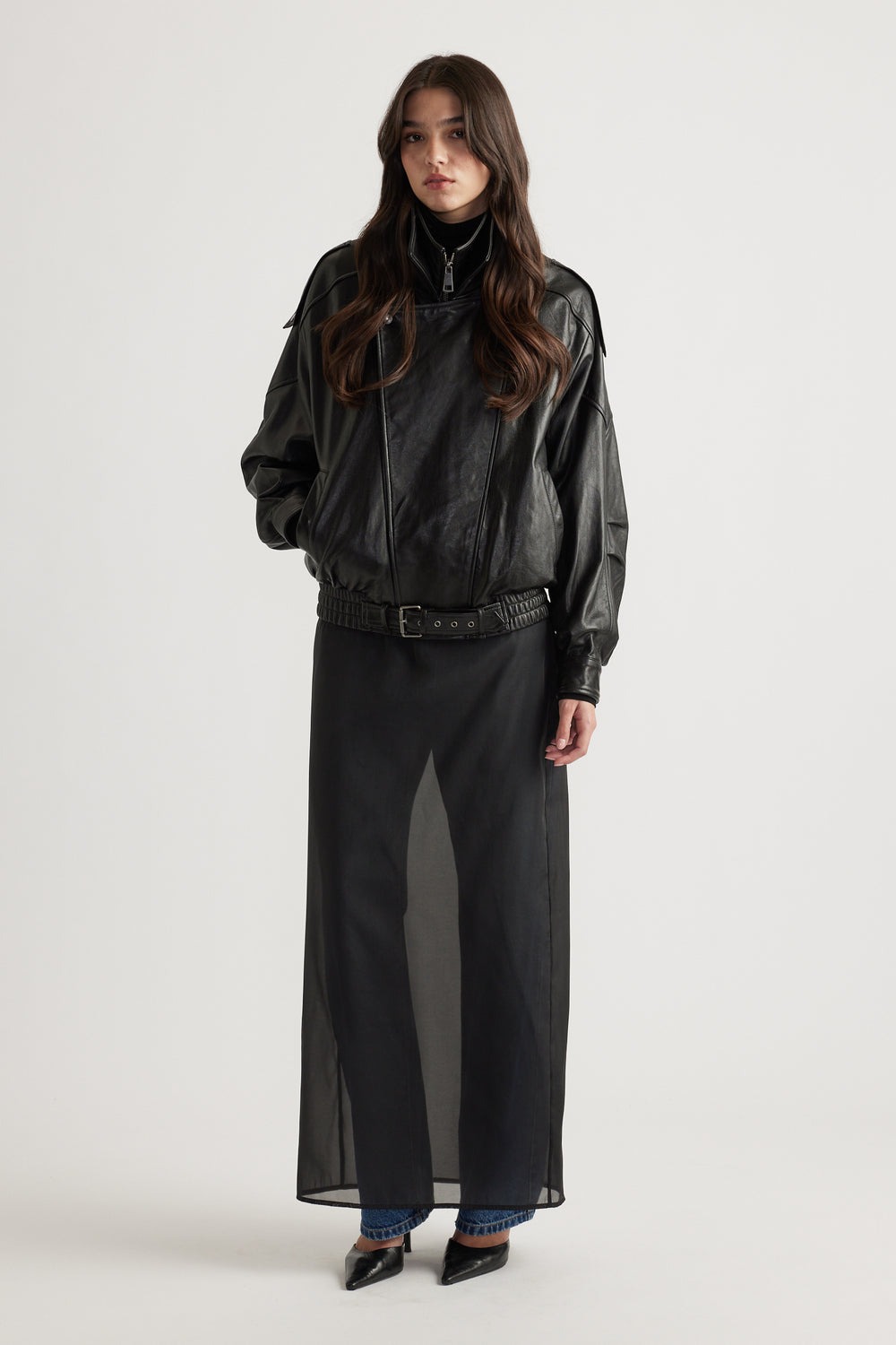 Young woman wearing an oversized black leather jacket layered over sheer black skirt and blue jeans with pointed black heels