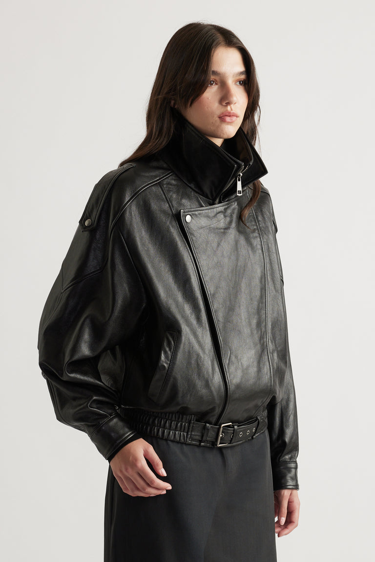 Woman wearing a black leather jacket with exaggerated collar and belted waist, styled with a black skirt