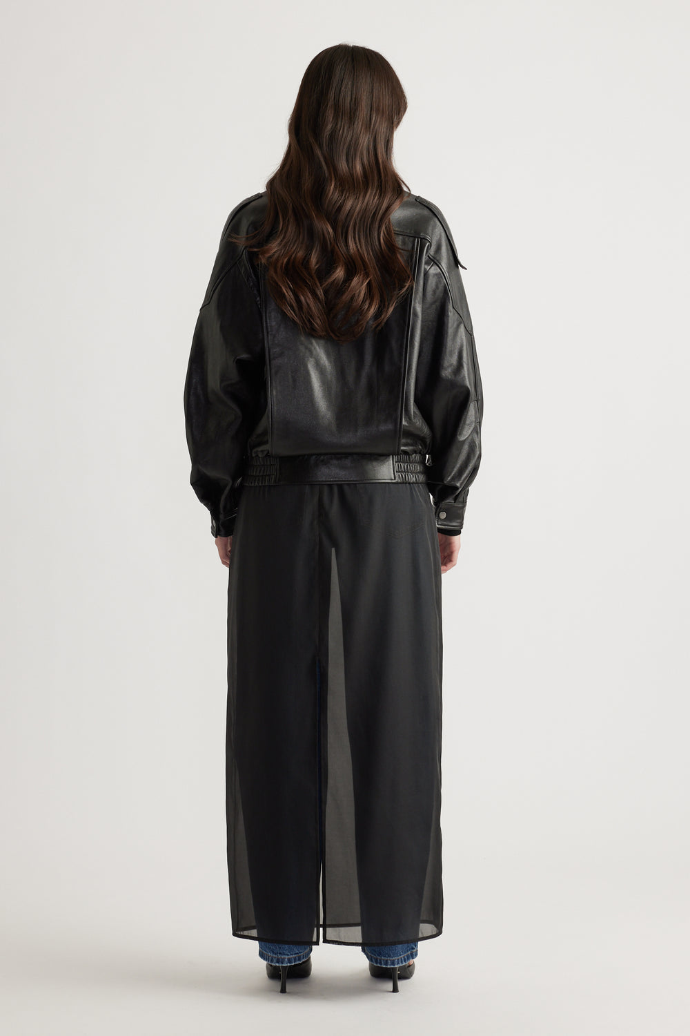 Woman seen from behind wearing a black leather jacket over a sheer long skirt with blue jeans and black heels