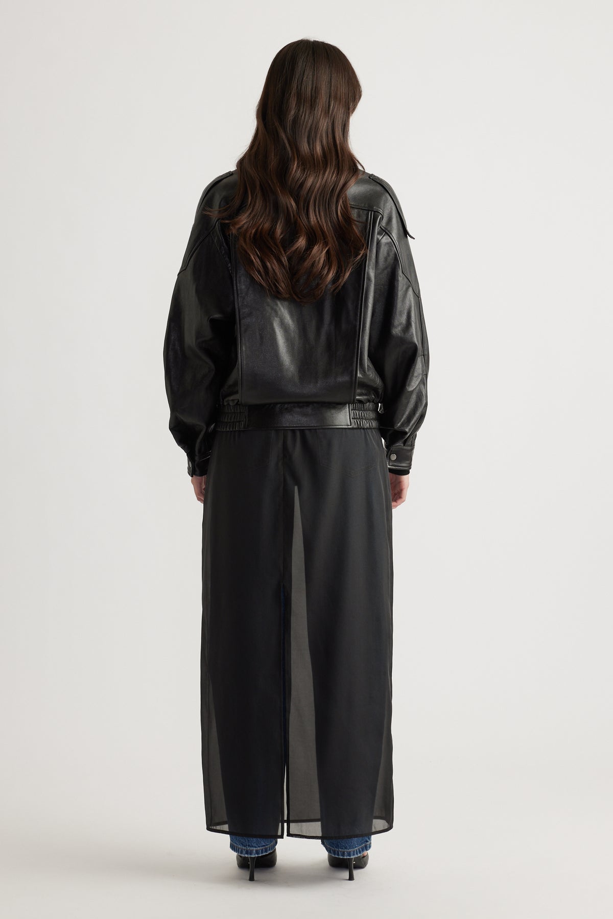 Woman seen from behind wearing a black leather jacket over a sheer long skirt with blue jeans and black heels