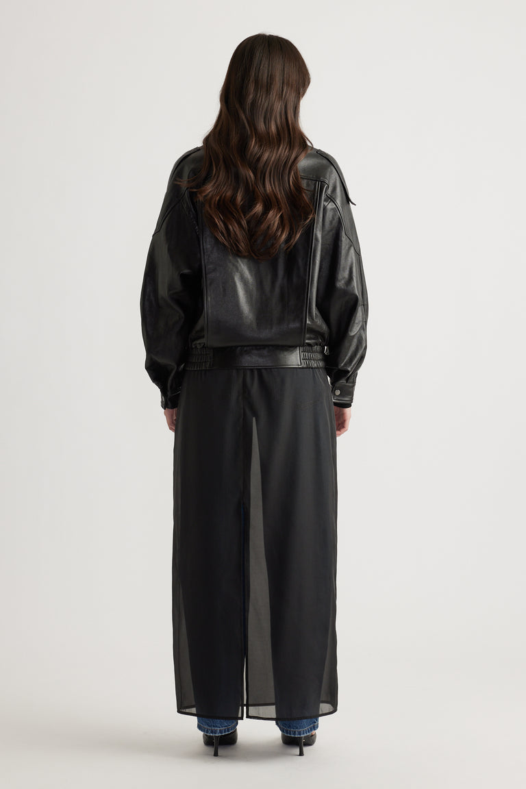 Woman seen from behind wearing a black leather jacket over a sheer long skirt with blue jeans and black heels