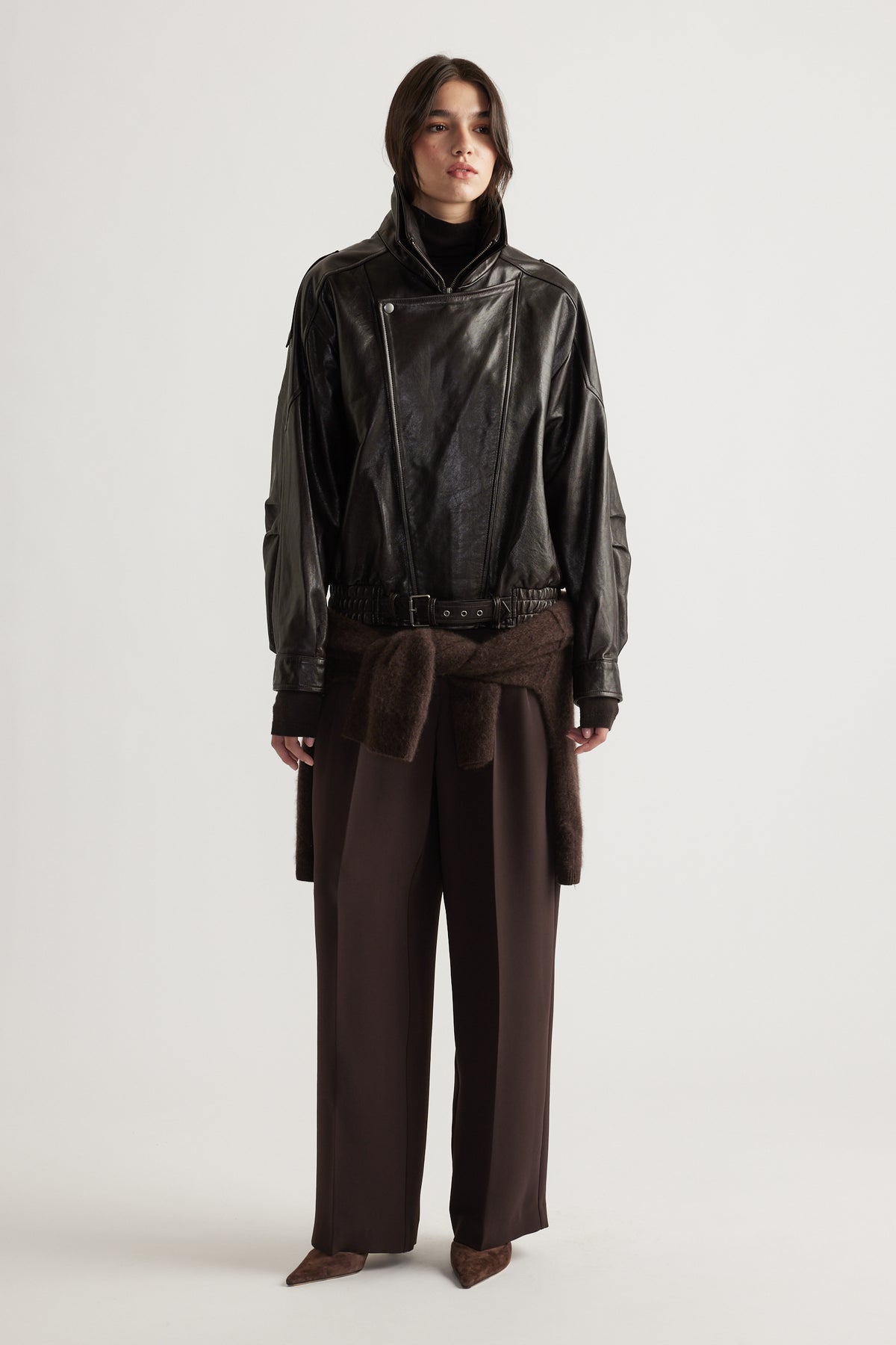 Woman wearing a belted black leather jacket with wide lapels paired with loose brown trousers and pointed shoes