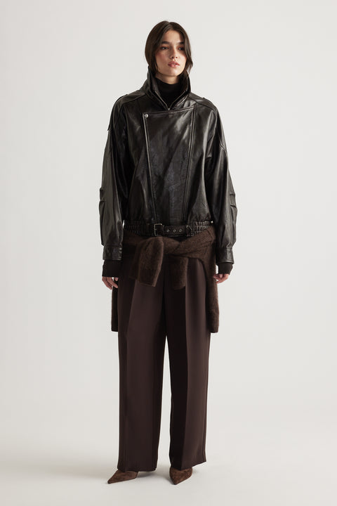 Woman wearing a belted black leather jacket with wide lapels paired with loose brown trousers and pointed shoes