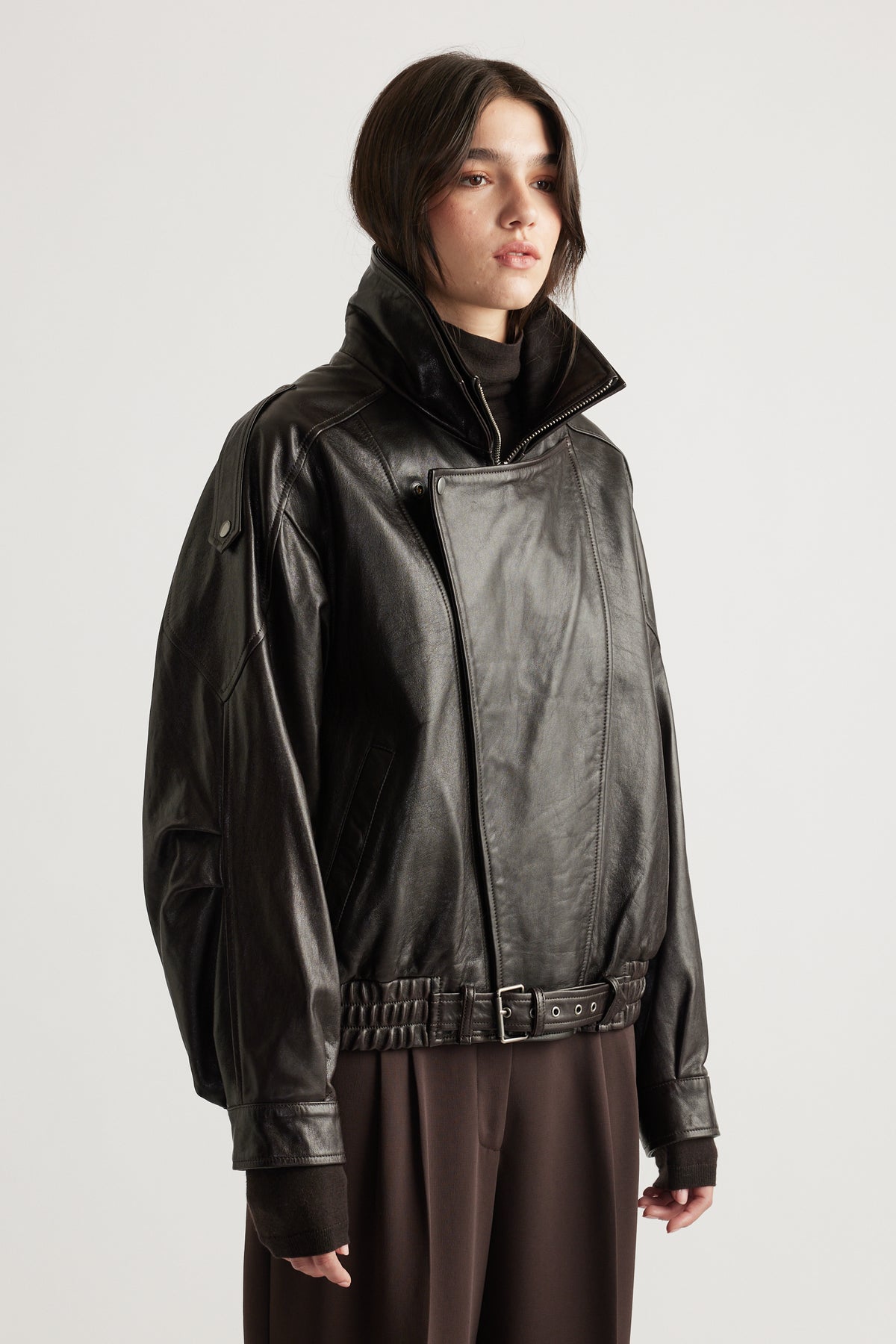 Woman wearing an oversized black leather jacket with high collar and belted hem paired with wide-leg brown trousers