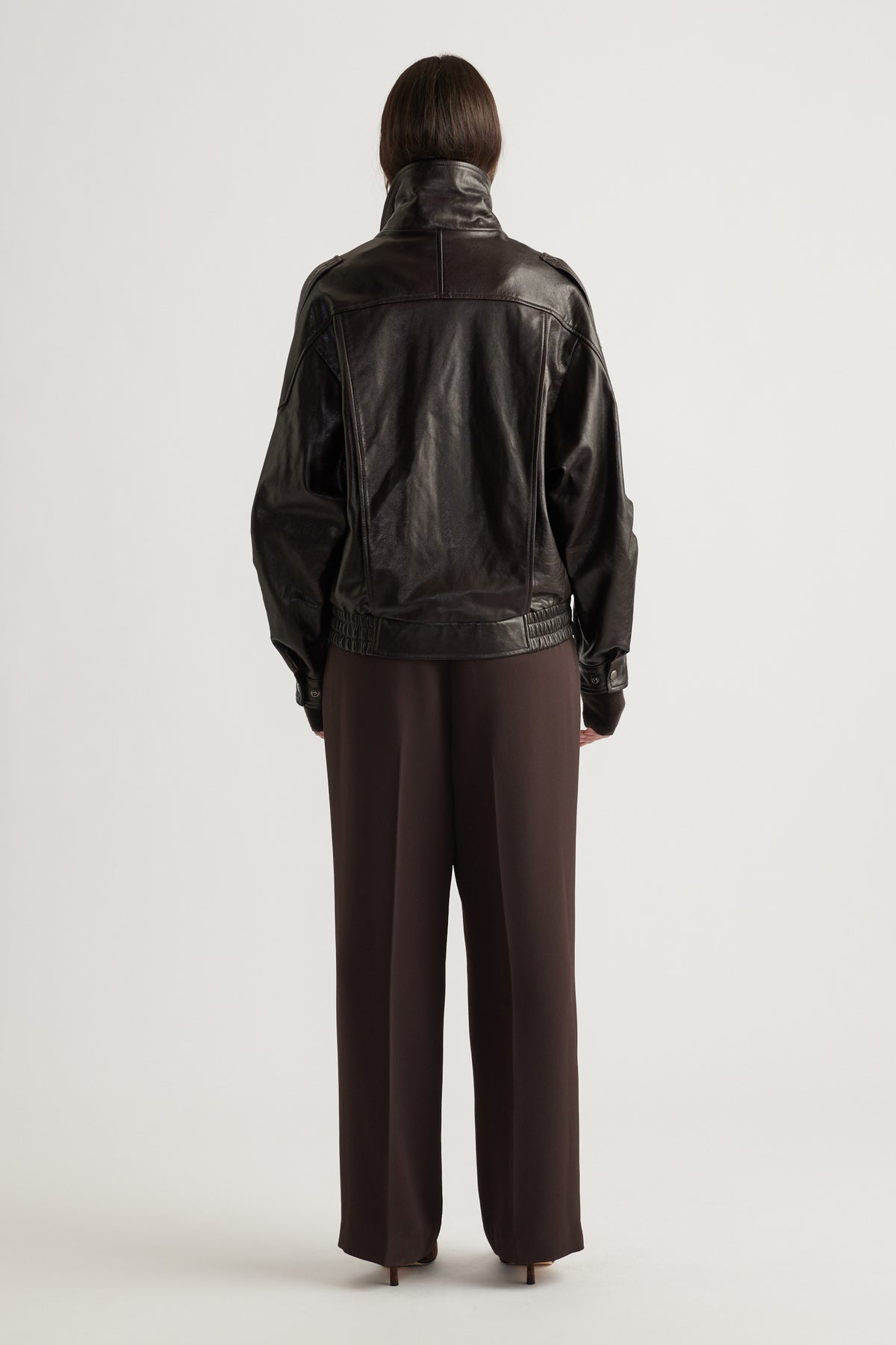 Rear view of a woman wearing a dark leather jacket with elasticated hem and straight brown wide-leg pants