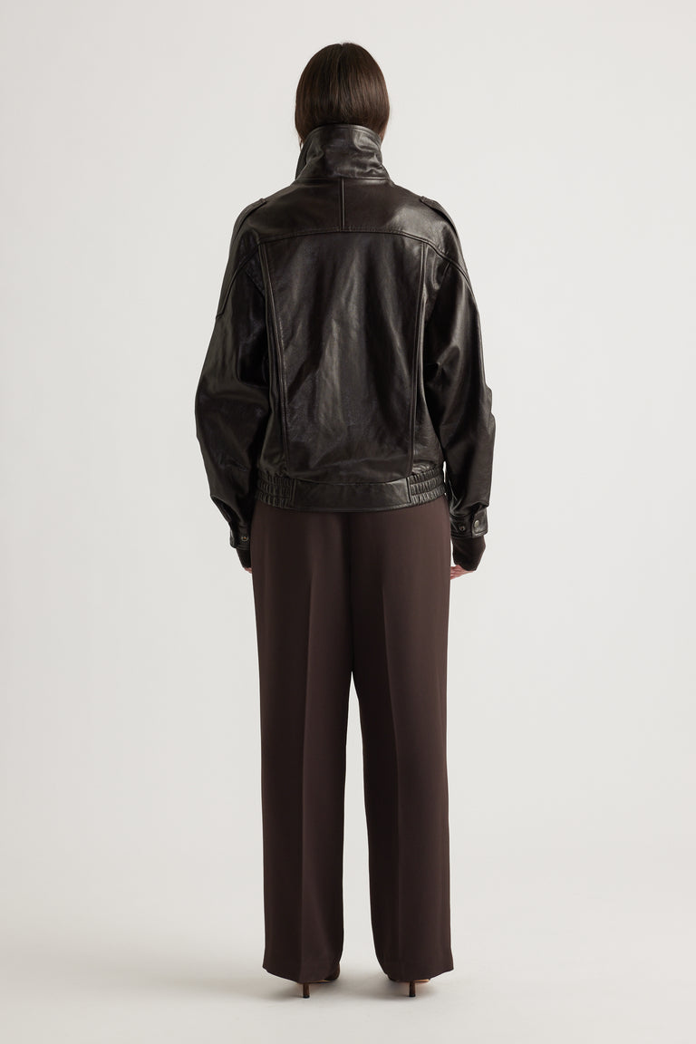Rear view of a woman wearing a dark leather jacket with elasticated hem and straight brown wide-leg pants