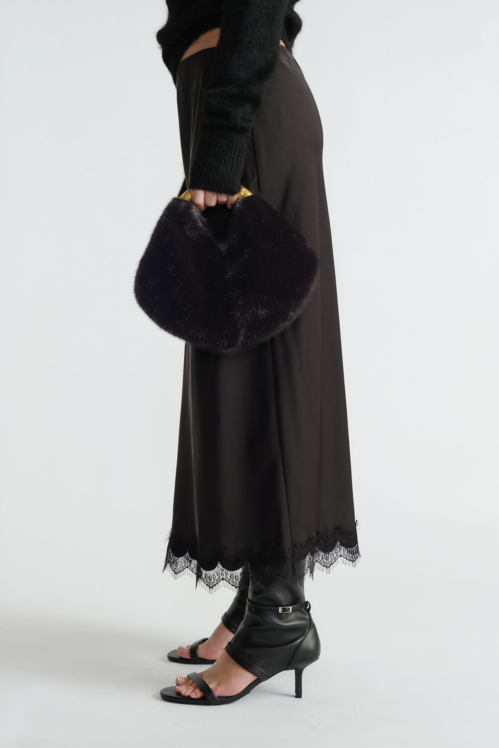 Woman holding a black faux fur bag with a gold metal handle, styled with a midi skirt featuring lace trim and black heeled sandals.