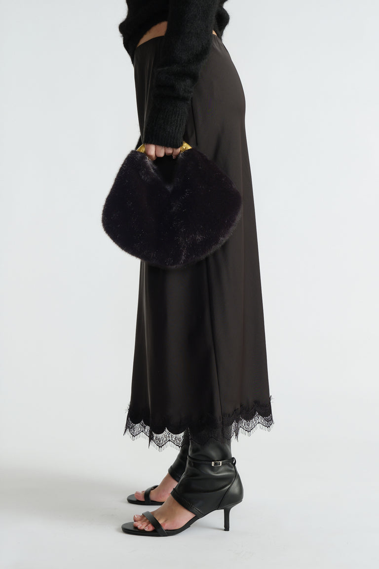 Woman holding a black faux fur bag with a gold metal handle, styled with a midi skirt featuring lace trim and black heeled sandals.