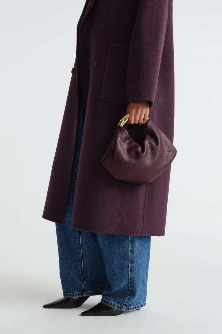 Deep plum leather bag with sculptural gold metal handle, styled with wide-leg jeans and a long coat.