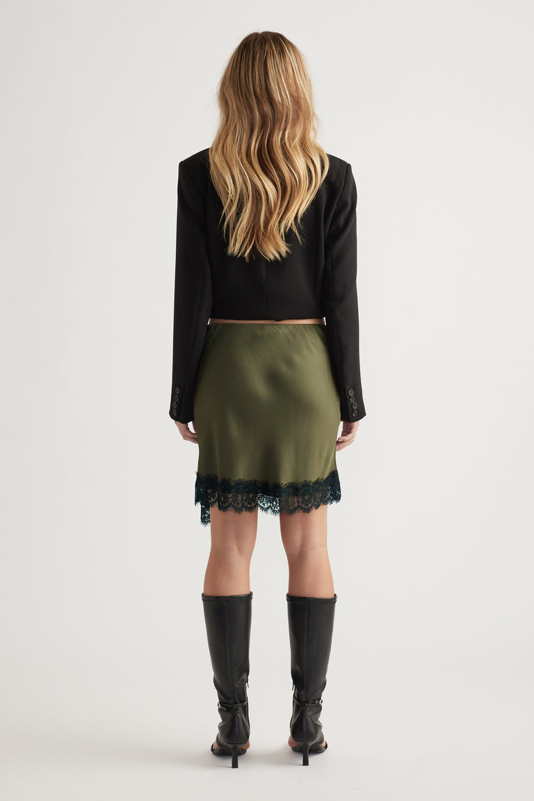 Woman wearing an olive mini skirt with black lace trim paired with a cropped black jacket and knee-high boots