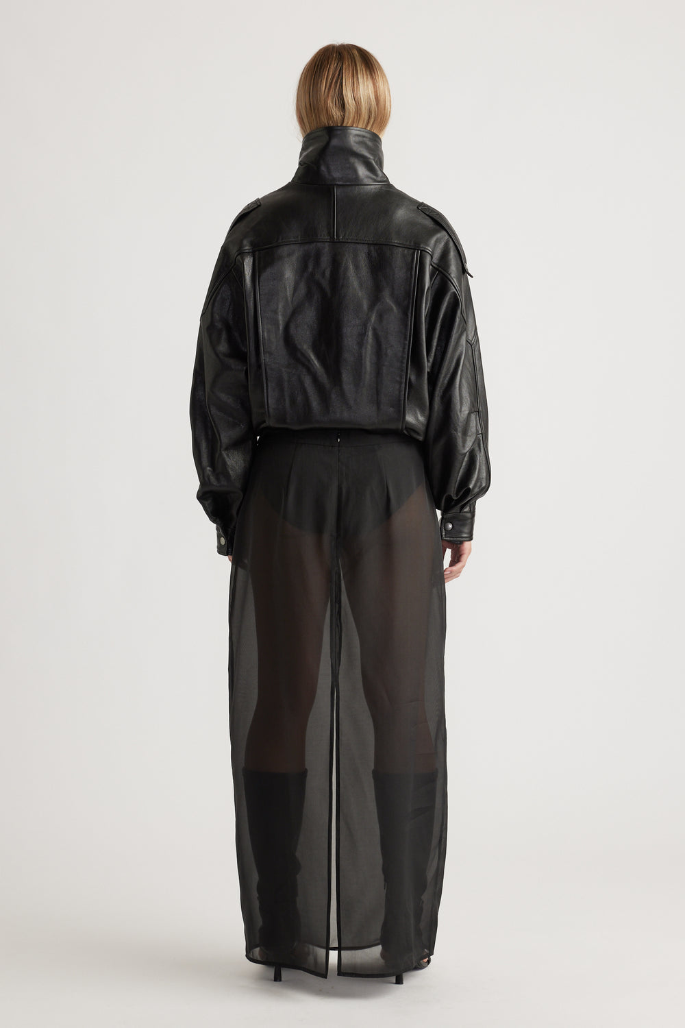 Back view of a woman wearing a cropped black leather jacket paired with a sheer black maxi skirt and heeled boots.