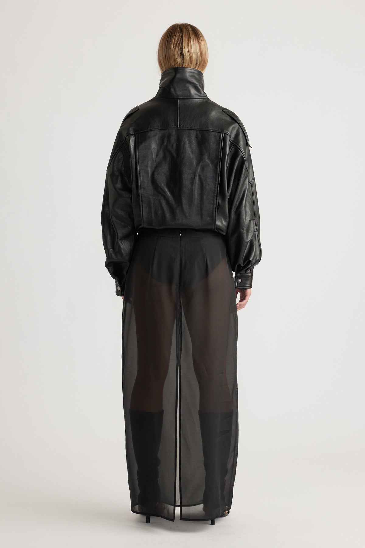 Back view of a woman wearing a cropped black leather jacket paired with a sheer black maxi skirt and heeled boots.