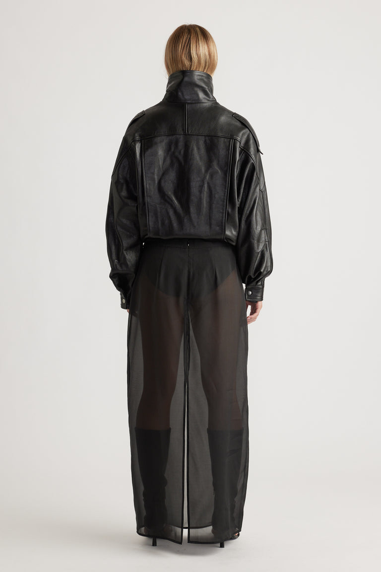 Back view of a woman wearing a cropped black leather jacket paired with a sheer black maxi skirt and heeled boots.