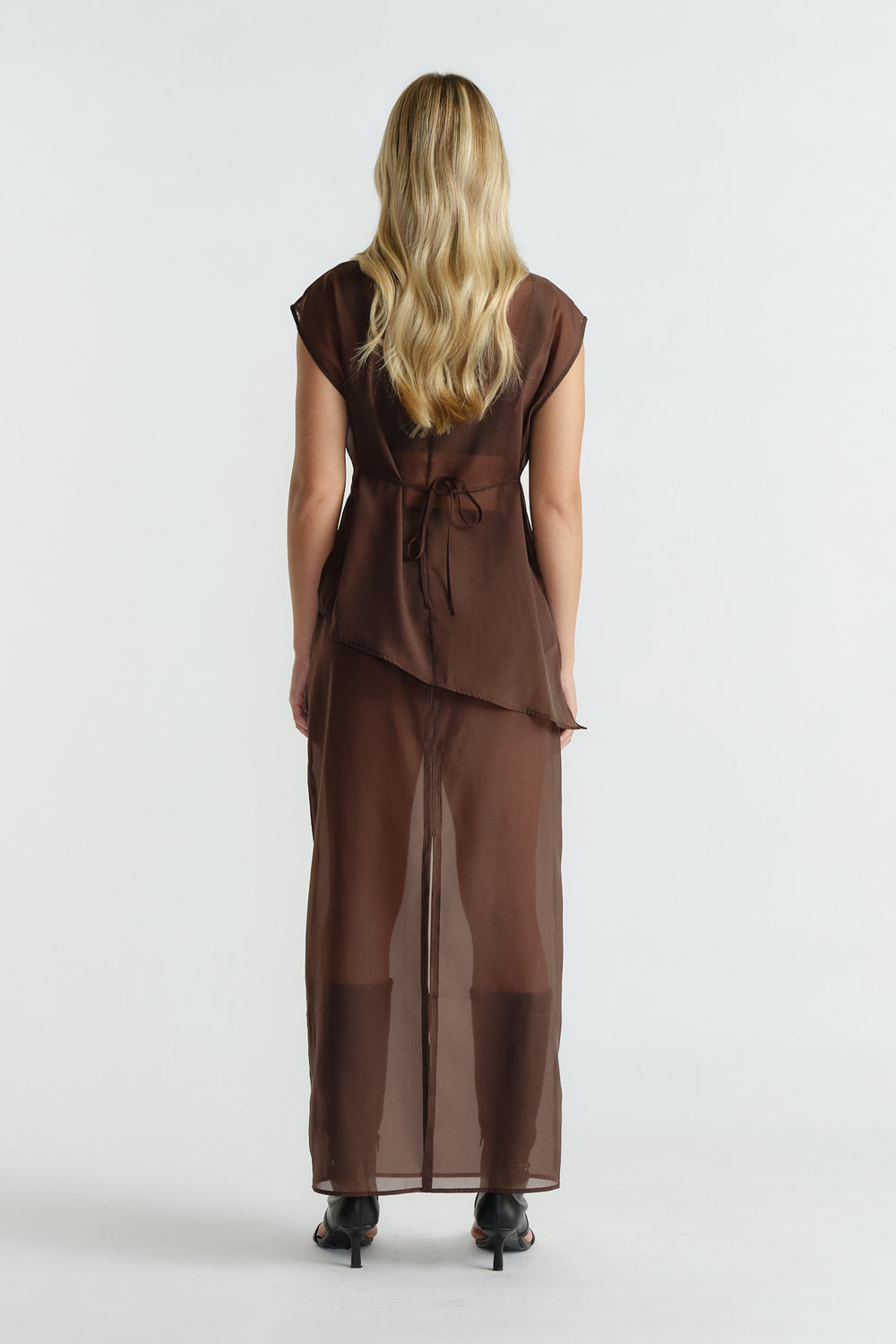 Rear view of a woman wearing a sheer chocolate brown maxi skirt and matching top with a tie-back detail and black heels