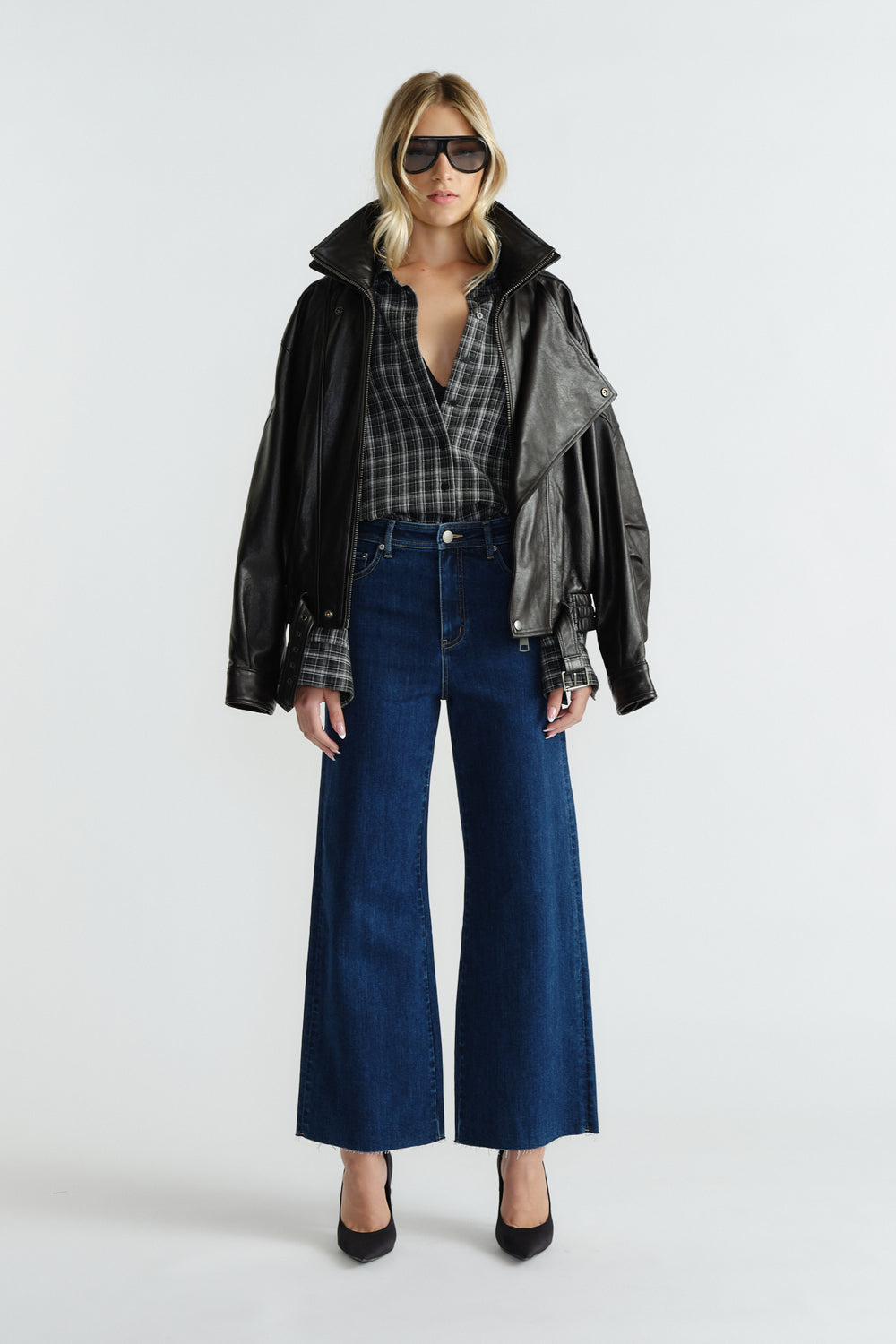 Woman wearing deep indigo wide-leg jeans styled with a relaxed plaid shirt, black leather jacket, and sunglasses