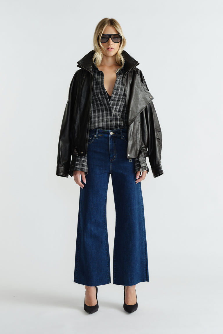 Woman wearing deep indigo wide-leg jeans styled with a relaxed plaid shirt, black leather jacket, and sunglasses