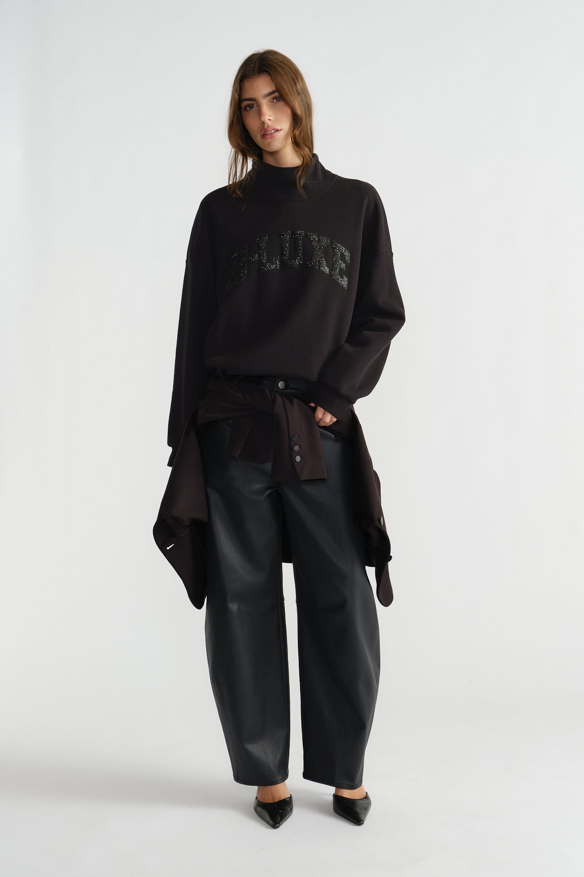 Woman wearing a relaxed black mock neck sweat with subtle sparkle detail, styled with loose black trousers and pointed shoes.