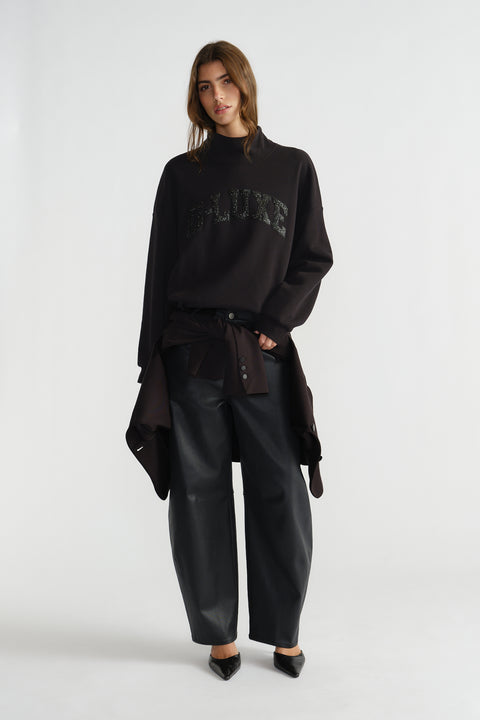 Woman wearing a relaxed black mock neck sweat with subtle sparkle detail, styled with loose black trousers and pointed shoes.