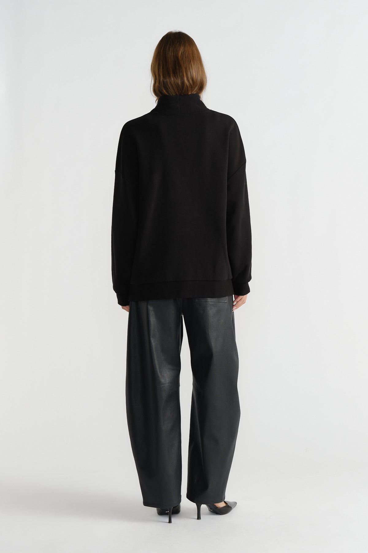 Woman wearing a black mock neck sweatshirt paired with wide-leg faux leather trousers and black heels, back view.