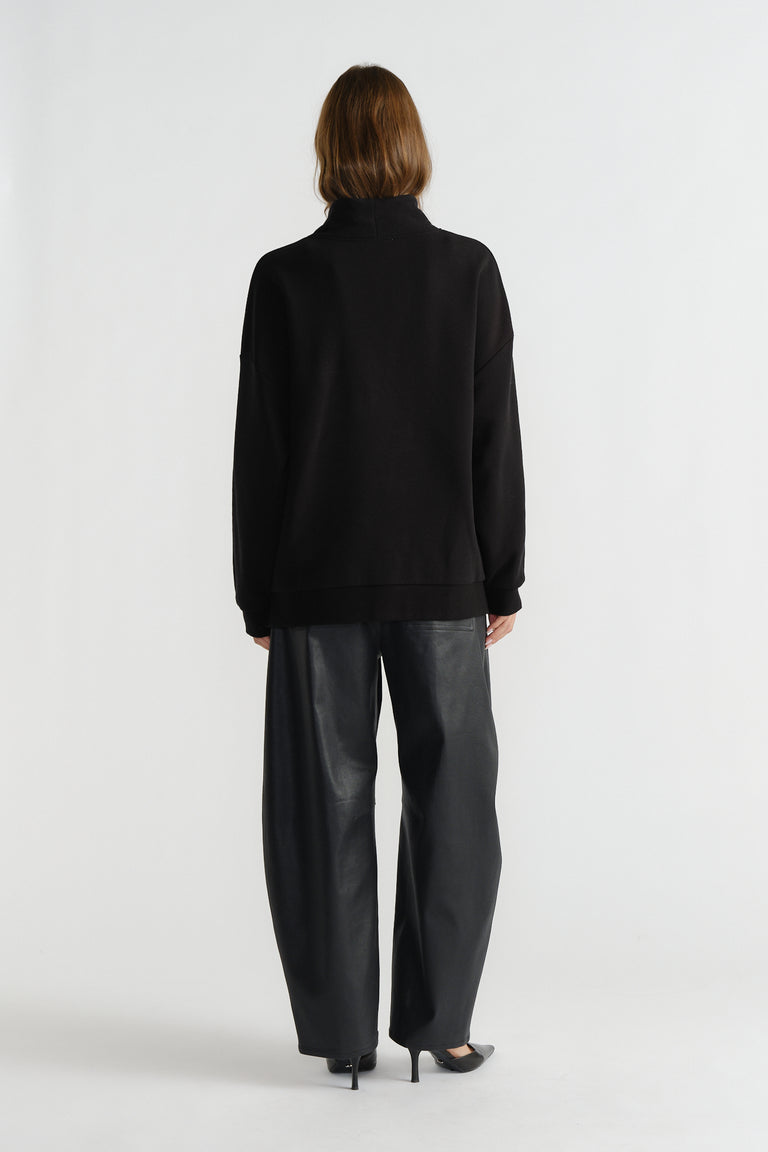 Woman wearing a black mock neck sweatshirt paired with wide-leg faux leather trousers and black heels, back view.