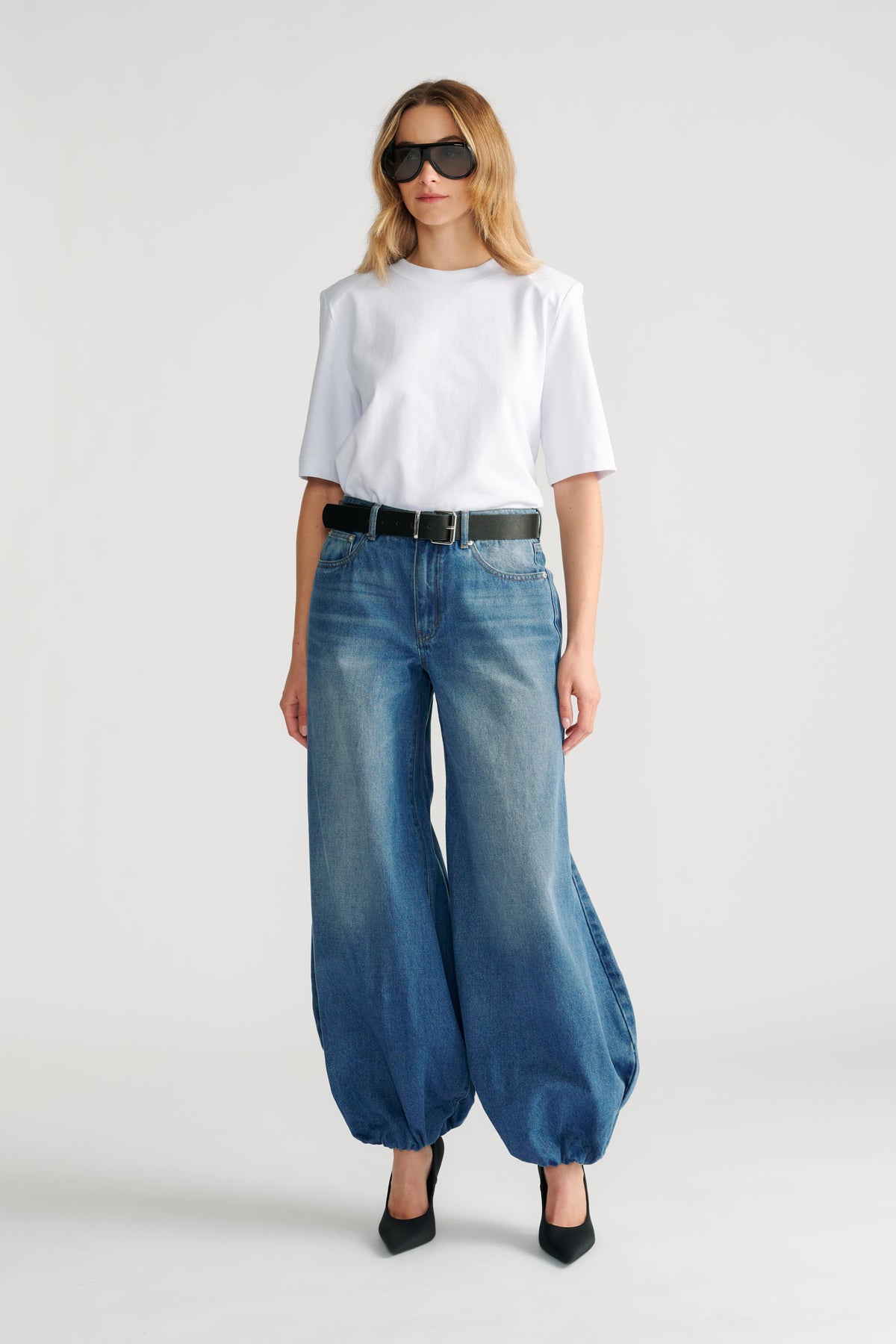 Woman wearing relaxed high-rise blue denim balloon jeans with a plain white tee, black belt, and pointed black heels.